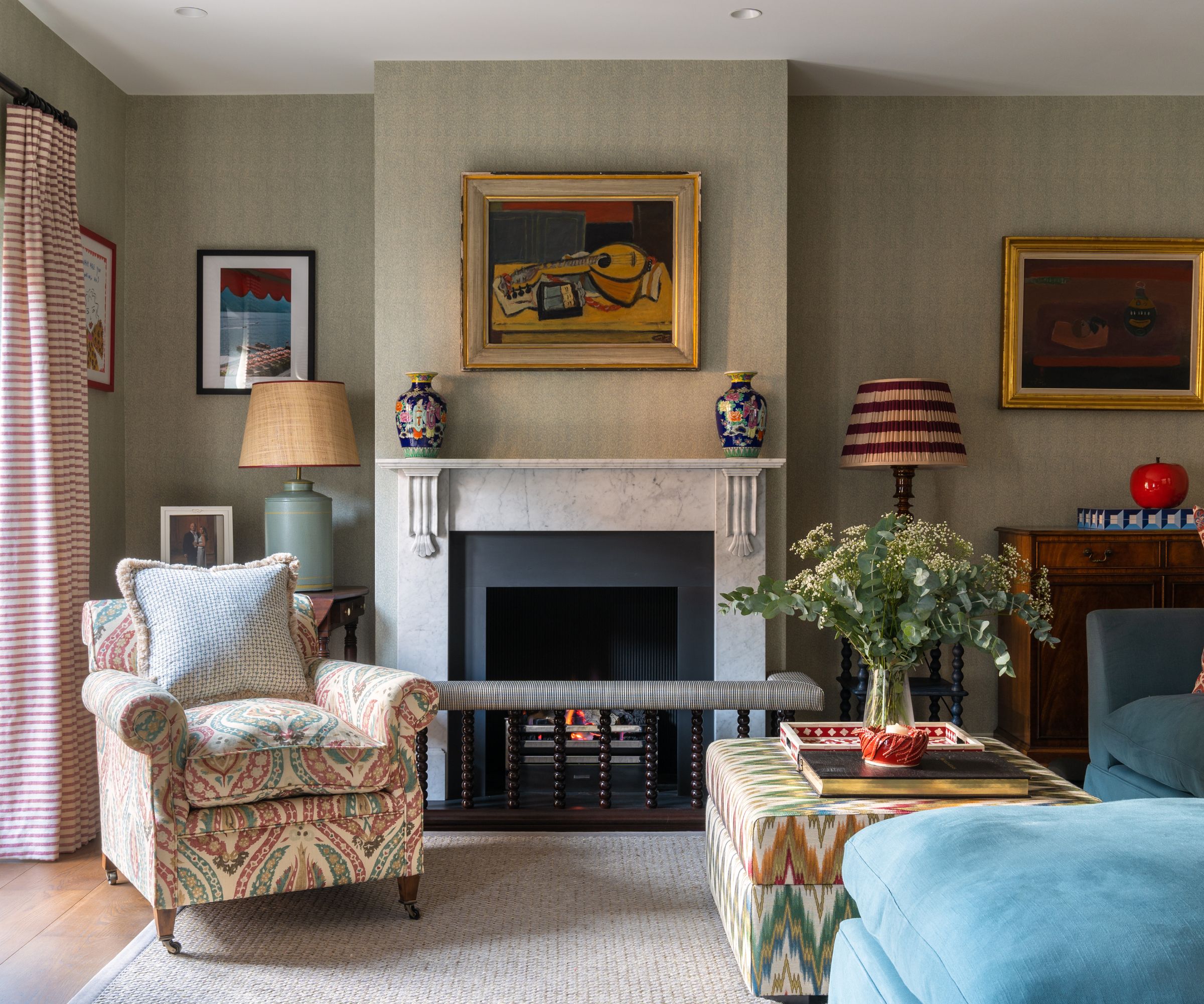 Salon avec art impressionniste au-dessus de la cheminée, un fauteuil rembourré, un foyer et un canapé en velours bleu et un pouf avec une plante