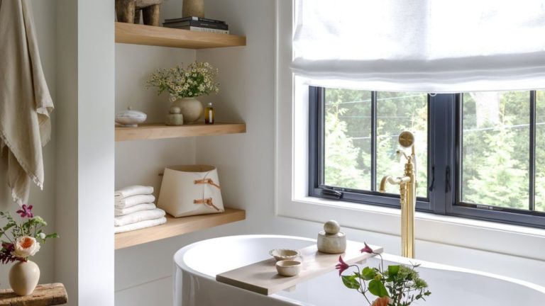 Bright white bathroom corner with deep bathtub, wooden shelves with neatly organized towels and bath products, and hanging towels. there are multiple vases of flowers in shot and a linen window treatment