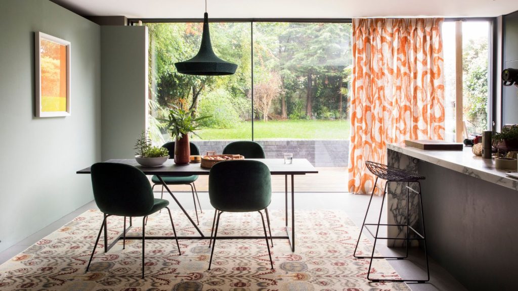 J’ai beaucoup d’opinions sur les aspirateurs, et oui, le vide V8 plus ancien de Dyson est toujours un excellent investissement en 2025 Dining room with table and chairs on patterned rug, picture window glass wall partially covered by patterned orange curtains, leading into and revealing patio garden.