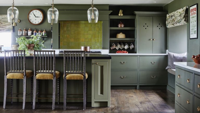 Dark green and brown kitchen area with kitchen island, high cushioned stools, hardwood floors, large glass lampshades and light green tiles.