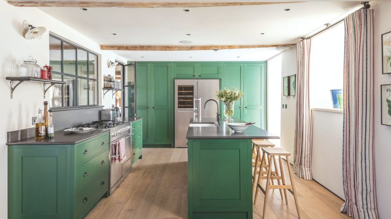 La « méthode de désencombrement de la fourche » peut sembler idiote, mais il est efficace pour apporter une simplicité à votre maison – et cela ne prend qu’un après-midi Kitchen and dining area with green cabinets, beamed ceilings, paneled window above gas range, kitchen island with faucet and flower vase next to high stools. On hardwood floors in well-lit room.