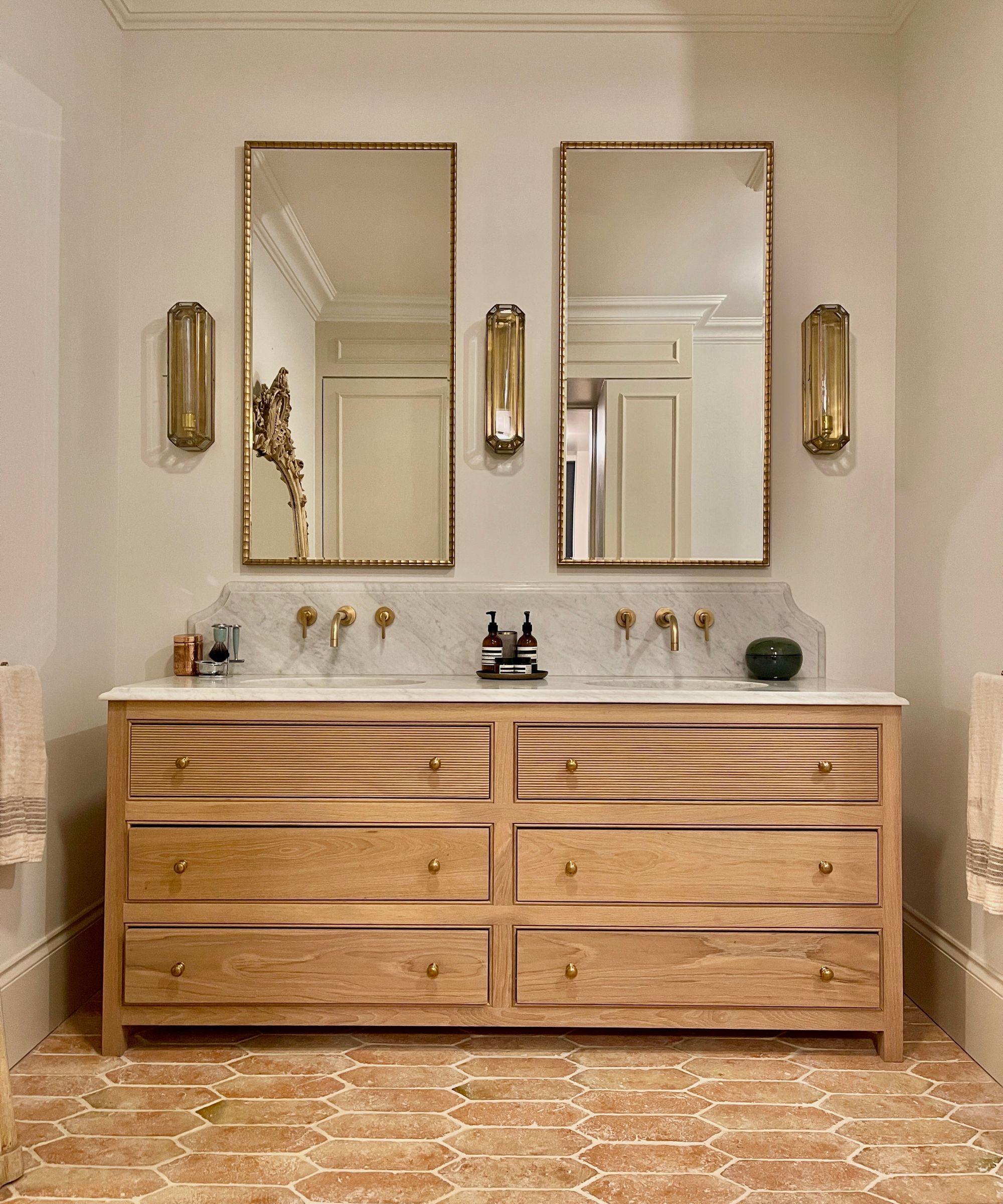 Une salle de bain classique avec une vanité en bois, des miroirs en or, un éclairage en or et des carreaux de sol en terre cuite