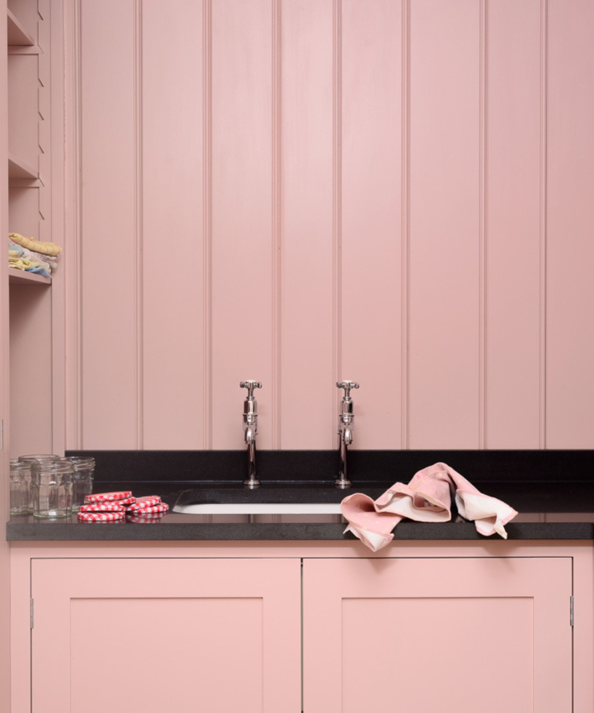 Société utilitaire rose avec une concentration sur l'espace de l'évier, qui a un comptoir en pierre noire, des robinets de chrome jumeaux et des étagères intégrées à gauche. Le mur derrière est lambrissée et les armoires, les murs et les étagères sont toutes une couleur rose bébé.