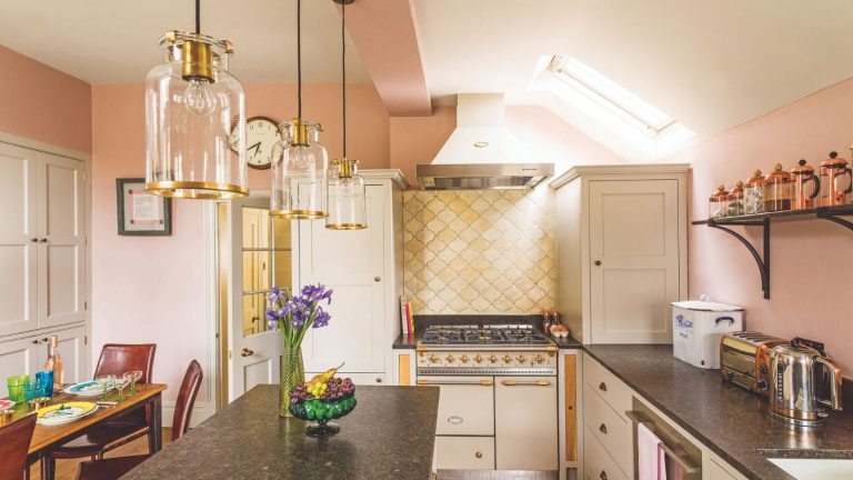 J’adore ce piratage super abordable et intelligent pour descendre ma bouilloire – cela empêche aussi les déchets alimentaires A pink kitchen with marble countertop, gold tiling, and glass and gold pendant lights. To the left is a wooden dining table with burgundy leather chairs.