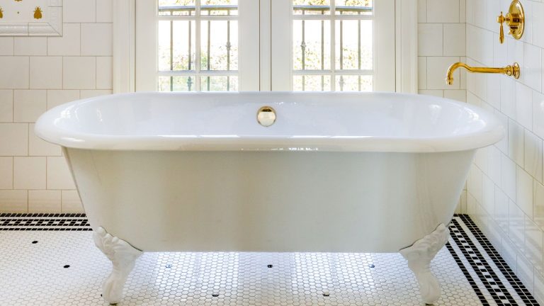 «Le gagnant des mains» – les experts du nettoyage réglent le débat intemporel: le rouleau de toilette face ou sorti? Spoiler: une façon est plus hygiénique White roll-top freestanding bath with gold faucet on wall. The floor is tiled white with black geometric border. The walls having white subway tiles and there is a balcony door feature in the background