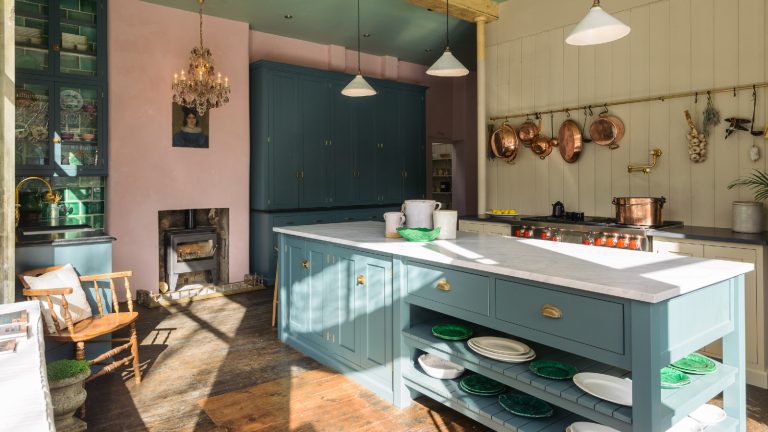 5 idées de stockage de bricolage que vous ne saviez pas que vous pouviez créer vous-même – ils sont aussi bons que sur mesure pour une fraction du coût kitchen with pink wall with stove, teal cabinetry, cream shiplap wall with brass hanging rail and copper saucepans, large kitchen island painted in teal, white countertop, wooden floorboards, alcove storage with glazed doors, wooden chair, chandelier and enamel pendant lights