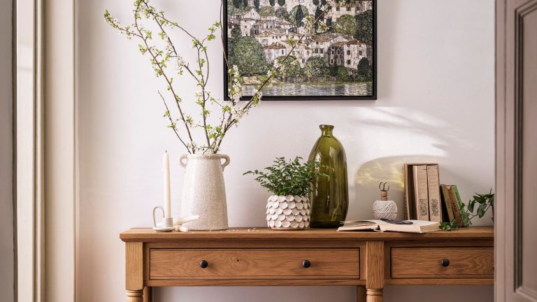 7 choses à désencombrer en mai 2025 – Définissez votre espace pour le succès de l’été avec des clairseurs simples et localisés A long wooden entryway table in a cream painted entryway. A cream vase with green stems in on top, next to a small green fern in a white pot, and an empty green glass vase.