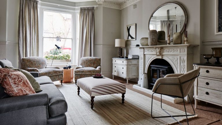 6 choses auxquelles faire attention lors de la réorganisation de votre maison pour éviter de faire la même erreur deux fois et de « refléter les changements de vie » Neutral living room with large white mantelpiece, circular mirror, white rug, and grey sofa. In the centre is a striped footrest. There are large bay windows looking over a street behind in the back.