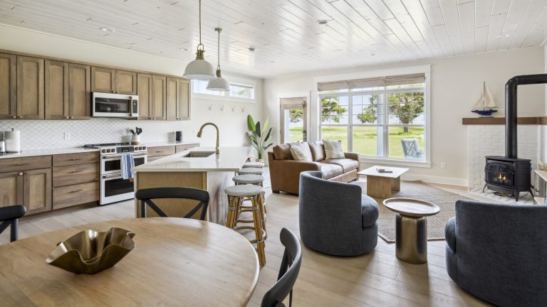 Pourquoi le Memorial Day sera le parfait – et peut-être la dernière fois – pour battre des randonnées tarifaires américaines pour les appareils de ménage à gros caisse Large open-plan space with kitchen island and stools; kitchen area with oven/stove cooker, wooden cabinets and microwave; living room area with sofa, armchair and small coffee table; large, round, wooden table in the foreground. All on wooden floor with bright windows in background.