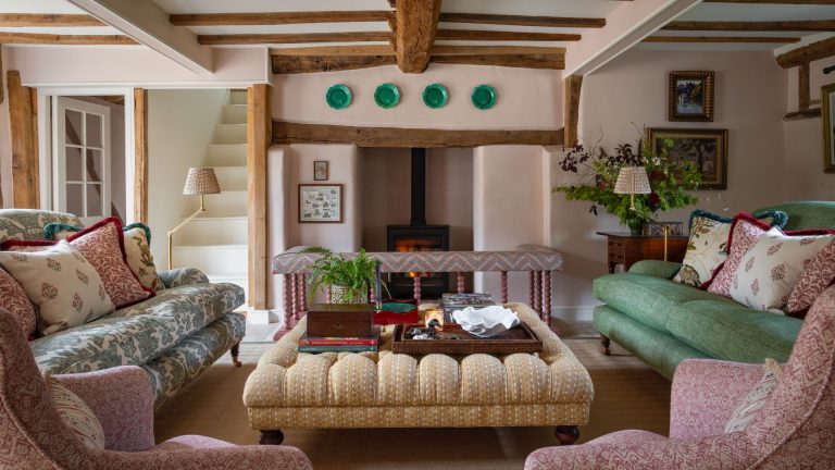 Devez-vous choisir d’abord votre couleur de peinture ou vos meubles? Croyez-moi, il y a une bonne commande si vous voulez un schéma de cohésion A country living room with a wood burning stove, two sofas opposite one another and two pink patterned armchairs in the foreground. In the middle of the room is a large yellow upholstered ottoman with a tray on top filled with books and plants.