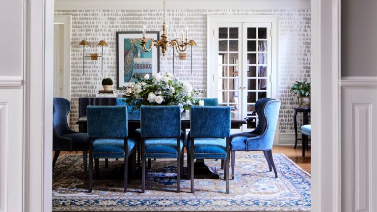 Une pandémie mondiale, une inondation massive et deux designers différents – ce projet, ses concepteurs et ses propriétaires se sont rendus en voyage pour se rendre à la maison familiale colorée que vous voyez maintenant Dining room with large dining table, blue velvet dining chairs, a gray and white wallpaper and Persian style rug