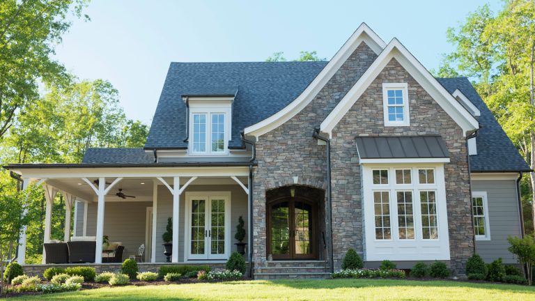 stone exterior of suburban house, with a porch wrapped around the left hand side. Surrounded by green trees.