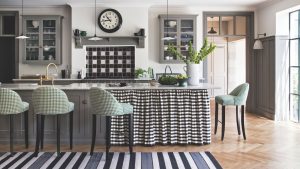 A black and white kitchen with grey cabinets and a wooden floor. In front of the black and white tiled hob is a marble island with a black and white gingham curtain, surrounded by green and white stools. In the foreground is a black, grey and white striped rug. Above the hob is a black clock, and to the right is an open doorway to a bright white room behind. There is a bright window to the far right.