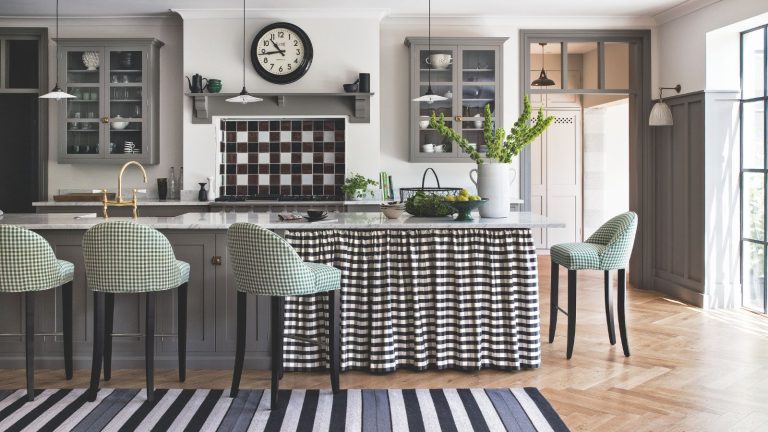 Embrasser le concept japonais de Kaji rendra un entretien ménager quotidien rapide, sans effort et établira un nouveau rythme pour les tâches A black and white kitchen with grey cabinets and a wooden floor. In front of the black and white tiled hob is a marble island with a black and white gingham curtain, surrounded by green and white stools. In the foreground is a black, grey and white striped rug. Above the hob is a black clock, and to the right is an open doorway to a bright white room behind. There is a bright window to the far right.