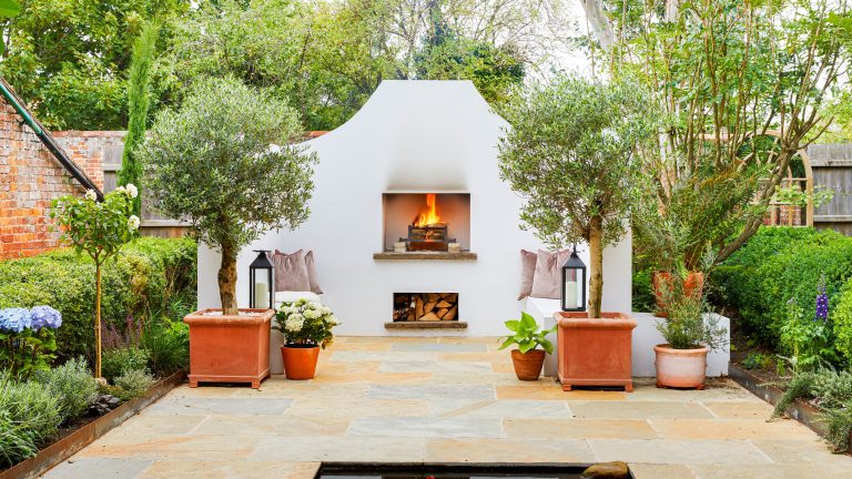 Secret hideaway Mediterranean style garden with white stone walls and seating, open fire place, log storage, grey cushions, flag stone patio with pond, lanterns, hydrangea and trees in terracotta planters