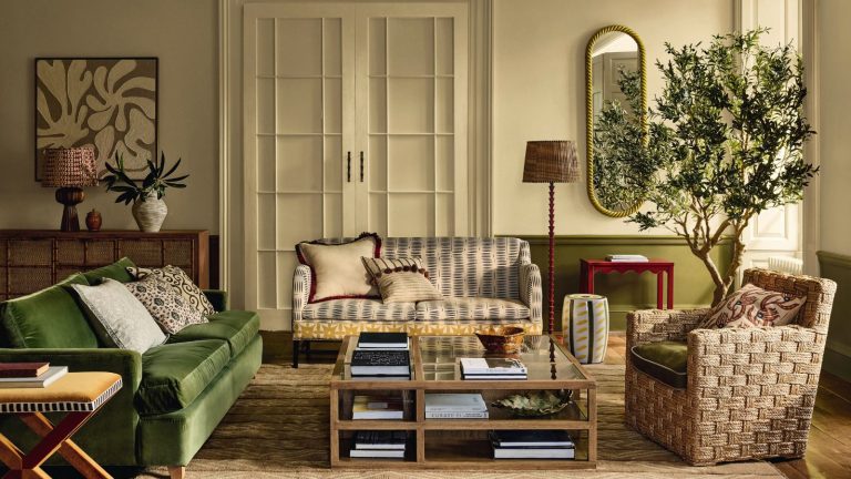 A warm toned living room with a green sofa, white patterned sofa, and a wicker chair. An olive tree in the background, and a glass square coffee table.
