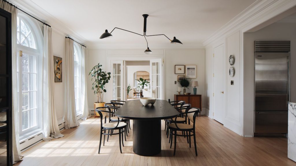 Neutral dining room with large dinner table and six chairs, abstract light, white walls and ceiling, two arched doorways and windows with open curtains, and a doorway leading to other room in background. To the right, a large fridge freezer is pictured.