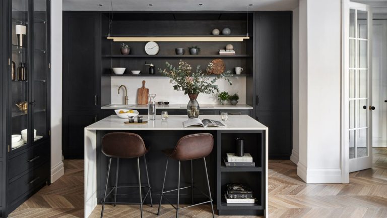 A black kitchen with wooden flooring, a white island with brown bar stools, and black shelving filled with bowls, chopping boards and a white clock. To the right is a glass and black cabinet and to the right is a white wall and white glass paned open door.