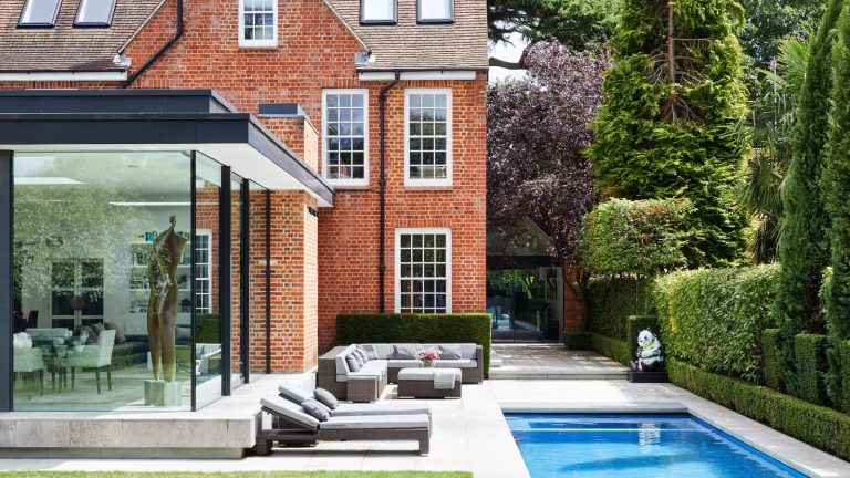 A red brick house and surrounding garden with a glass conservatory, pool, grey loungers and sofas, and leafy green bushes and trees.