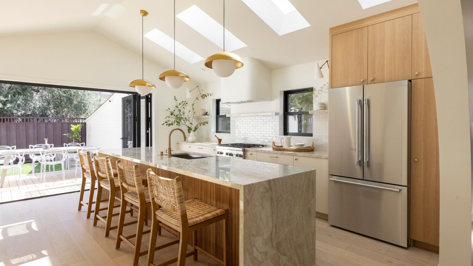 Cuisine moderne avec grand congélateur de réfrigérateur en acier inoxydable, îlot de cuisine en marbre avec quatre chaises en rotin, trois lumières suspendues dorées. Sur la photo, c'est la cuisinière, deux petites fenêtres, un mur d'éclaboussures de carreaux en briques blancs et une zone extérieure avec table et chaises.
