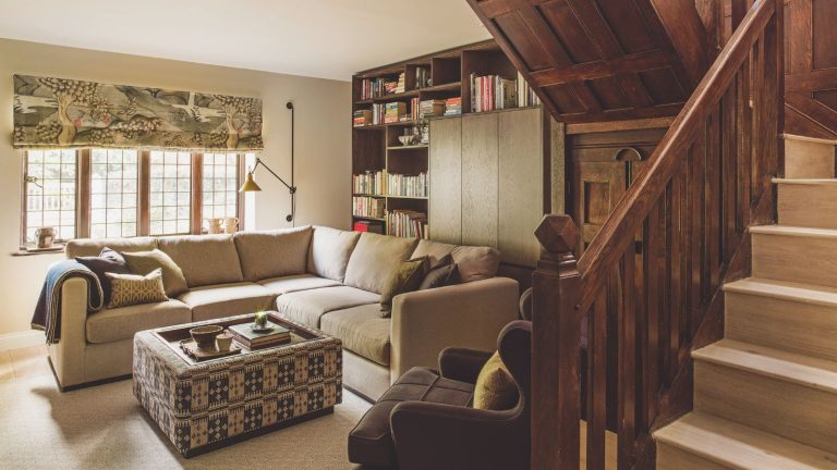 Small neutral living room with ornate wooden staircase and built in cabinetry at the far back wall. There is a patterned square coffee table in front of an L-shape sofa. It looks cozy but bright