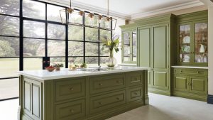 A large kitchen with green painted island and tall cabinets. Floor to ceiling windows behind.