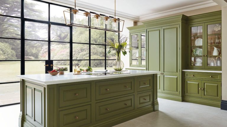 A large kitchen with green painted island and tall cabinets. Floor to ceiling windows behind.