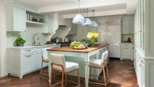 A midimalist kitchen with blue-green cabinets, terracotta floors, and a wooden countertop on the island