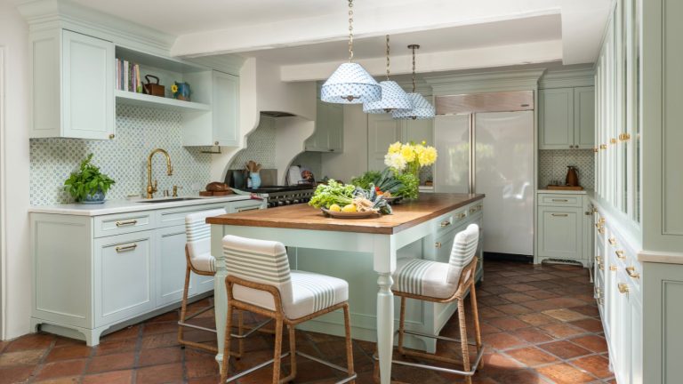 A midimalist kitchen with blue-green cabinets, terracotta floors, and a wooden countertop on the island