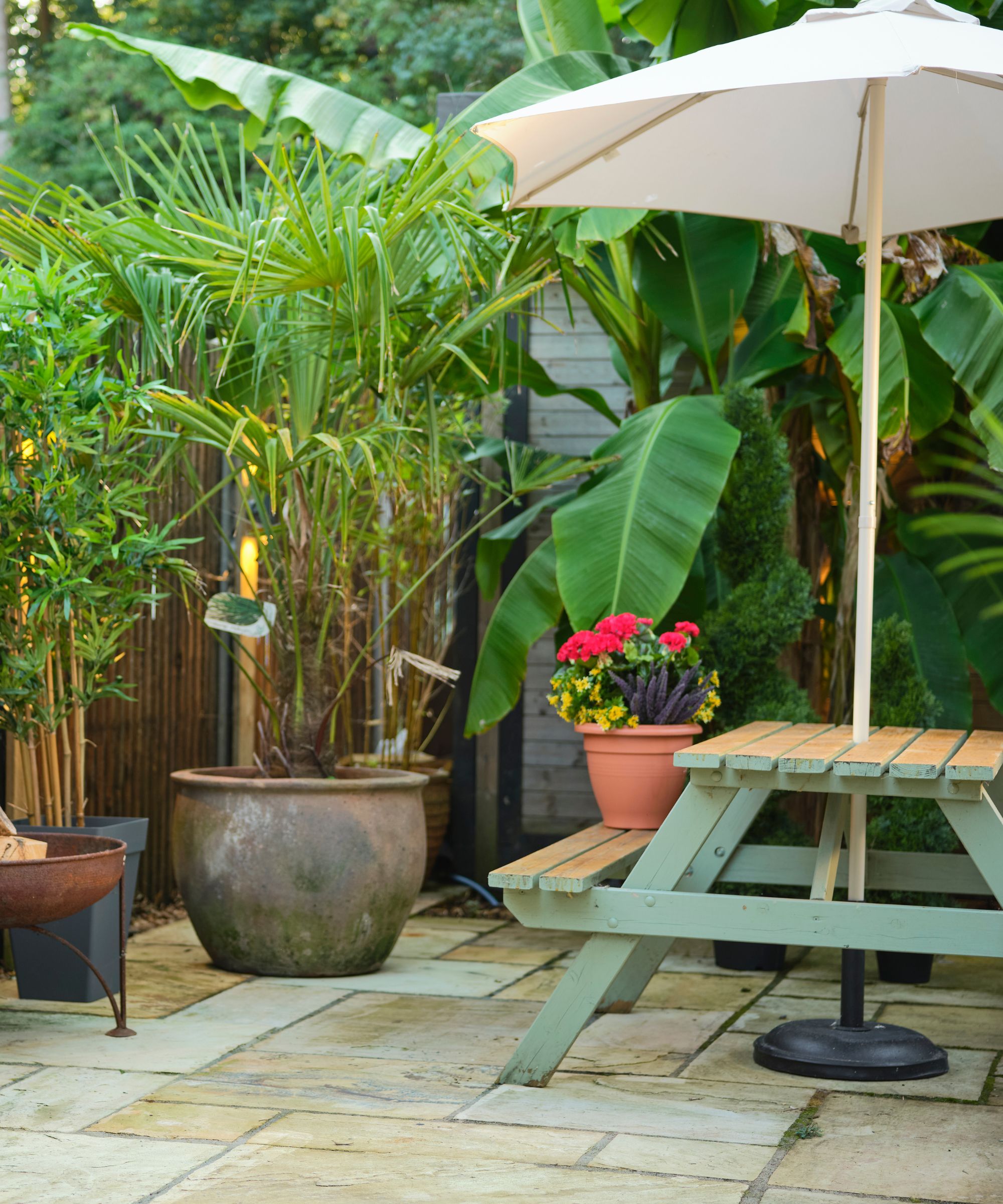 Jardin avec table verte et banc, grand parasol et plantes et verdure entourant la zone. Sur les planchers carrelés en plein jour.