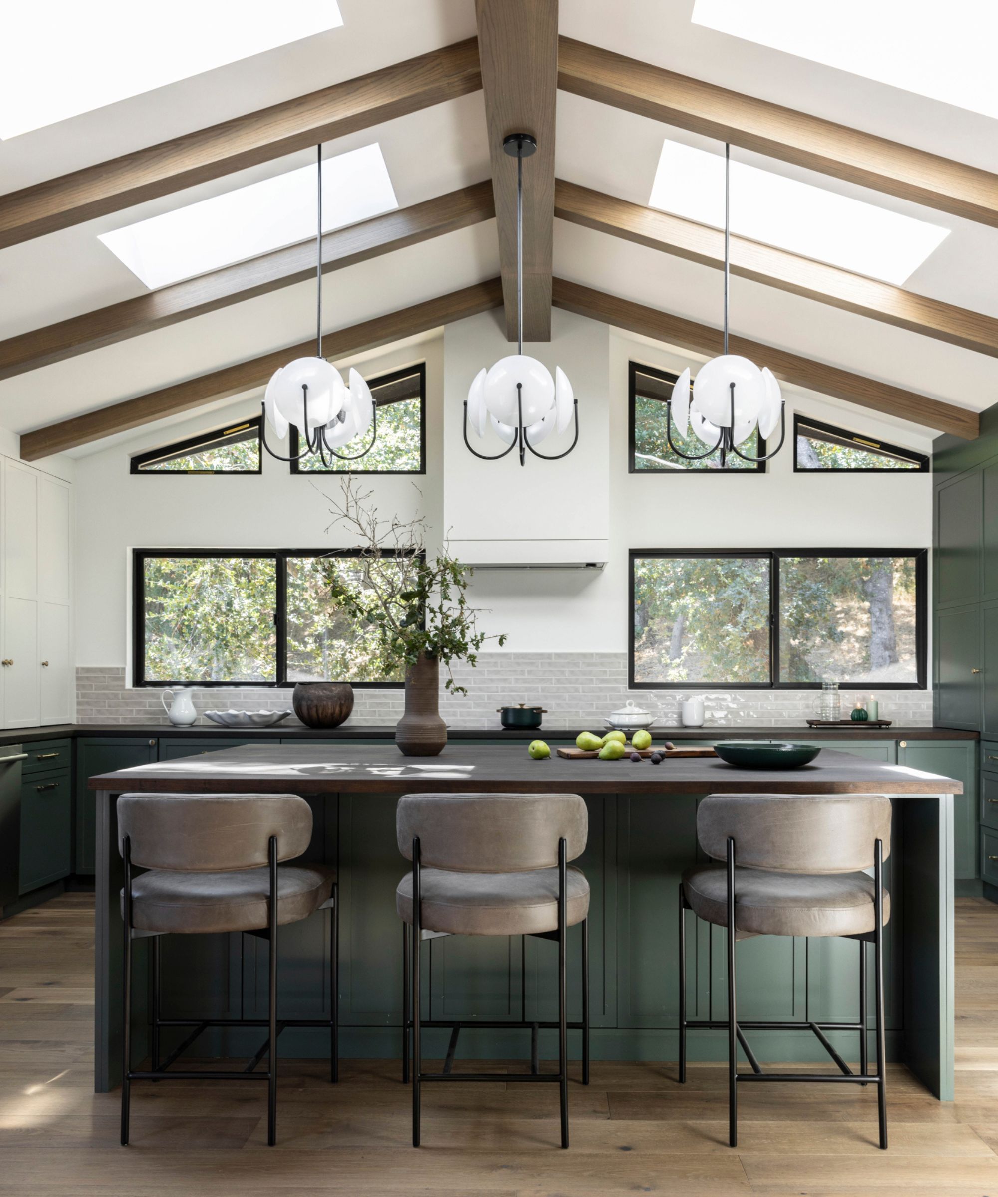 Cuisine lumineuse et moderne avec des lumières suspendues, îlot de cuisine et trois tabourets et plancher en bois. Sur la photo en arrière-plan, des fenêtres révélant un espace extérieur, un mur et une cuisine en briques et en briques