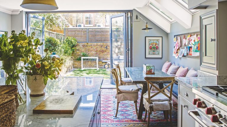 Bright kitchen and dining area with large plants in vases, kitchen island, dining table with cushioned chairs, skylights, and open doors leading to small, sunny, outdoor area.