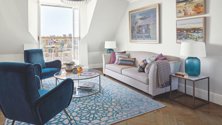7 choses à nettoyer en juin 2025 – Bienvenue à l’été dans un style désinfecté et éviter les problèmes de ravageurs A sunny living room with blue velvet chairs, a white and blue patterned rug, and a pale beige couch with colorful cushions. To the right of the couch is a marble and metal coffee table with a large blue lamp with a white shade, and above are framed painted artworks. In the centre of the room is a circular glass coffee table. Open doors onto a balcony are seen in the background.