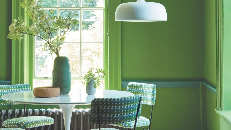 A bright green painting dining room with a white dining table, surrounded by green chairs. A white pendant hanging from the ceiling.