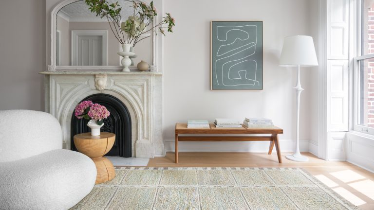 Ces 5 couleurs de tapis sont démodées pour 2025, selon des designers d’intérieur Living room with cream colored textured area rug, curved armchair, wooden side table and bench, fireplace with mirror above and floor lamp