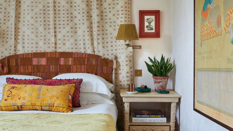 A maximalist bedroom. A bed with a red patchwork headboard, yellow bedding and pillows, and a patterned fabric wall hanging behind. A yellow pleated fabric lampshade on a wall lamp, above a light wooden bedside table.