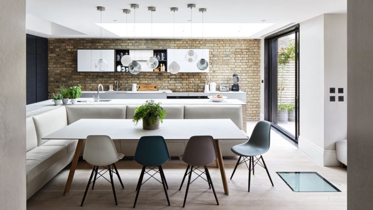 A bright and modern kitchen with an exposed brick wall, white cabinets, white kitchen island, white corner couch, white table and mismatched chairs. To the right is an open glass door.