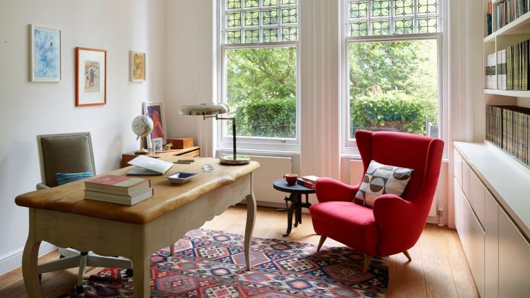 Juste un petit objet intelligent a doublé l’espace de stockage sur mon petit bureau WFH et aide à la douleur quotidienne au cou A bright home office with large windows, a wooden desk and a plush red armchair with patterned cushion. On the desk are piles of books and a table lamp. Behind it is a wheelie grey chair. On the wall are three framed artworks. On the opposite wall is a large bookshelf and white cabinet. The floor is wooden, with a patterned rug in the center.