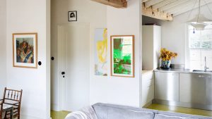 Des armoires en acier inoxydable, un éclairage rétro et un sol jaune vif ont transformé cette cuisine d’appartement fatiguée des années 1960 en l’espace d’inspiration parisienne les plus fraîches « sans effort » An open plan kitchen and living area with white walls, stainless steel cabinetry and art work on all of the walls