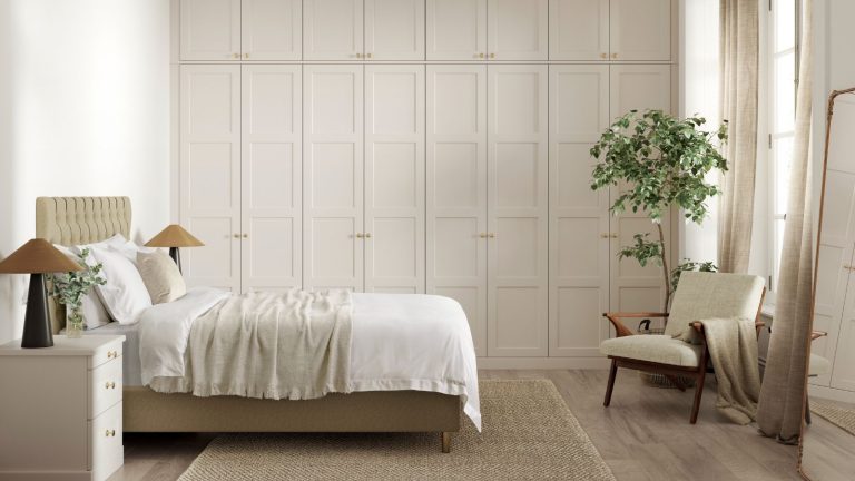 A neutral bedroom with large built in closets along the back wall with panelled walls. A double bed in front of them with two symmetrical night stands, a chair opposite