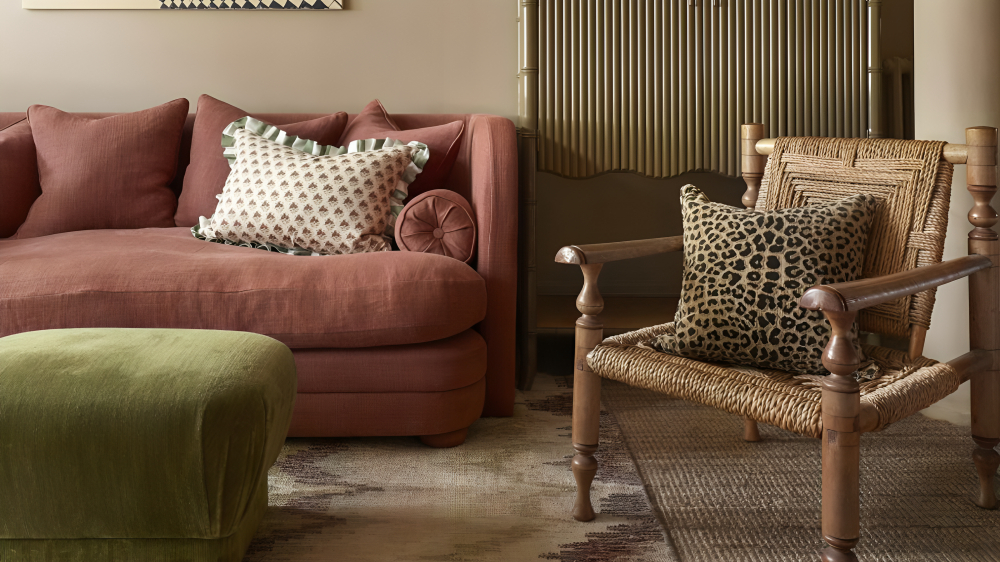 Un salon avec un canapé rose, un tabouret de tissu vert et une chaise à bras tissé avec un coussin imprimé léopard dessus.