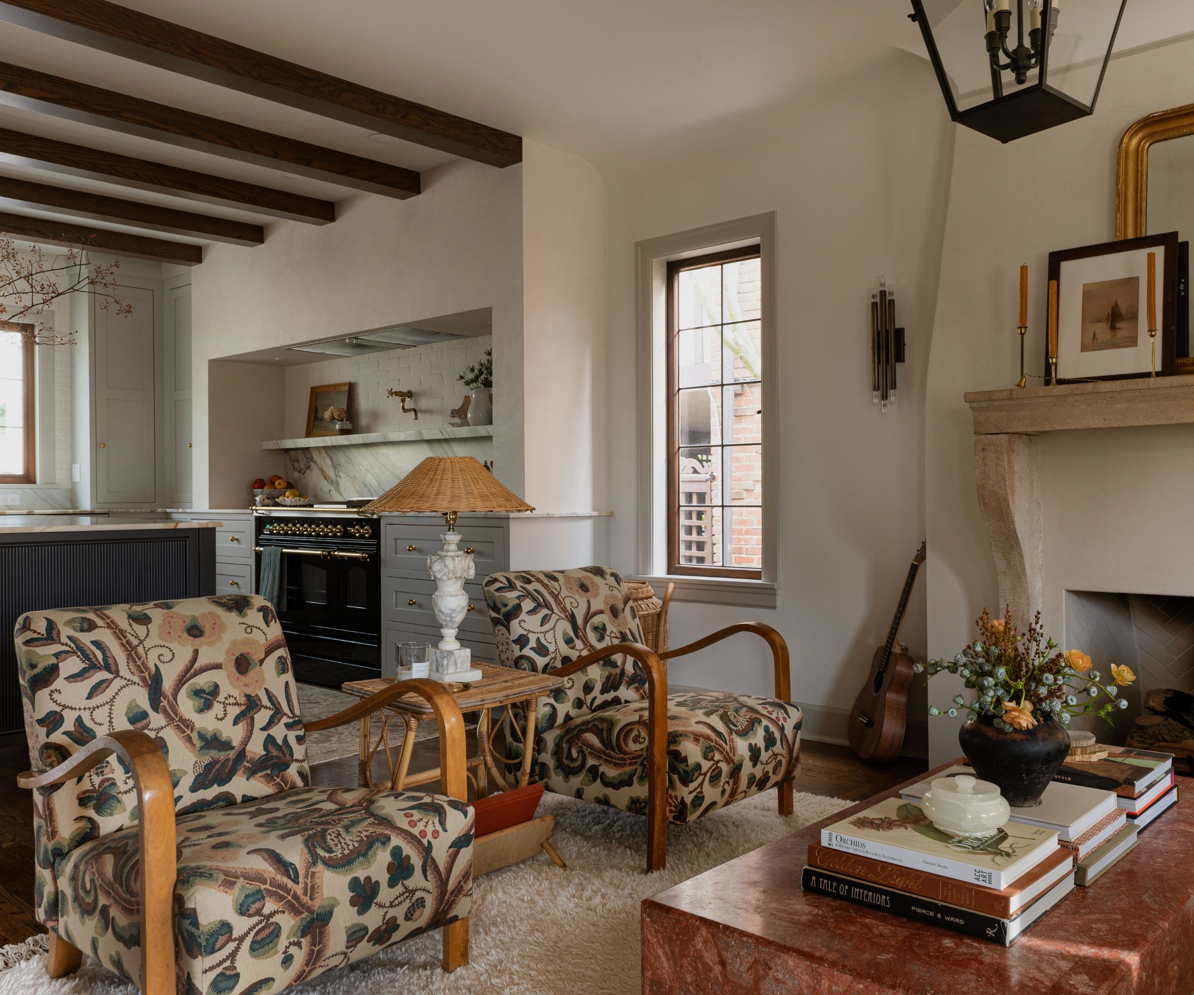 Salon dans une cuisine ouverte avec manteau en pierre, chaises vintage rembourrées dans une imprimé floral vintage, table basse en pierre décorée de livres de table basse