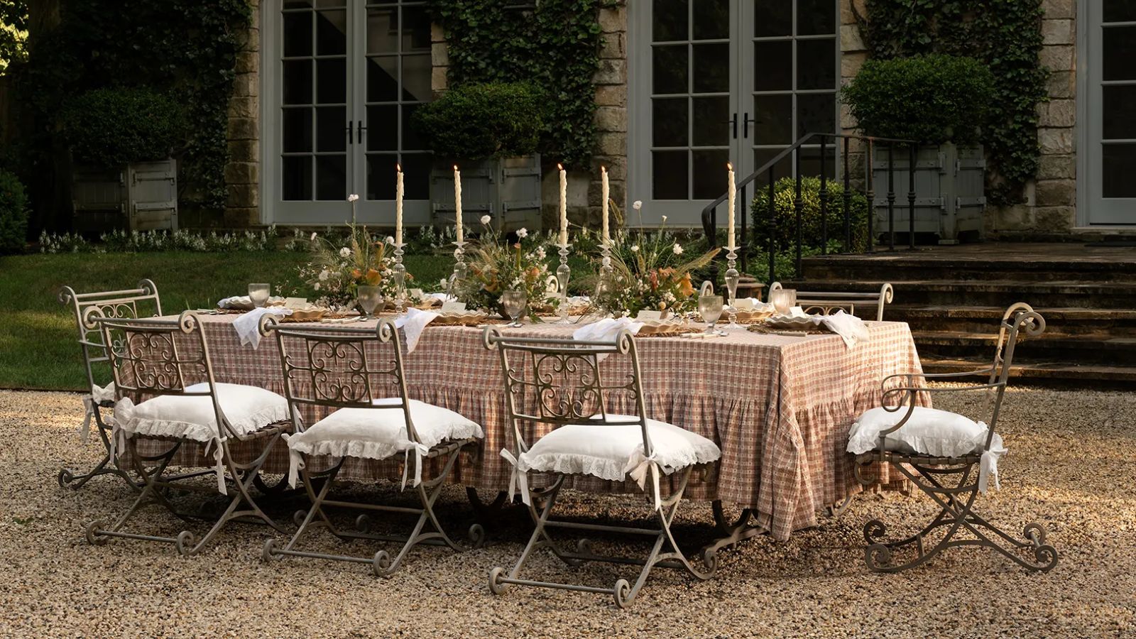 Grande table extérieure longue coiffée sur une terrasse de galets avec une nappe à volants à carreaux roses, des chaises ornées, posées de fleurs et de grandes bougies à cône