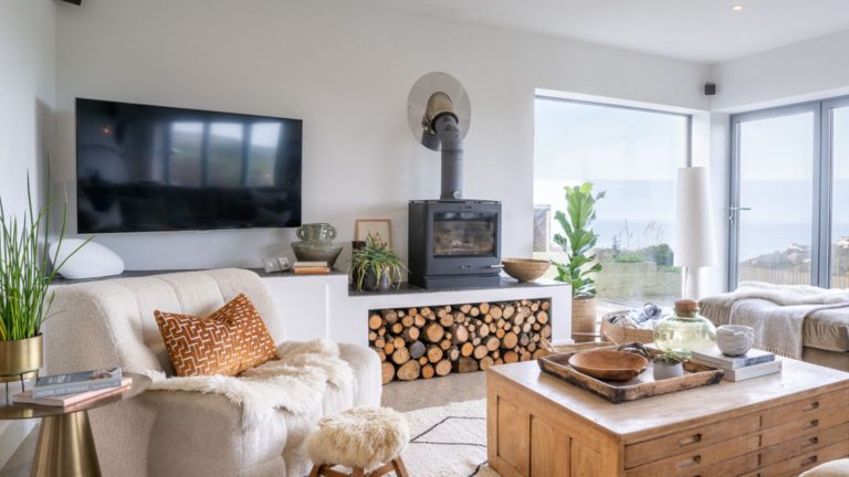 A neutral living room with a large wall-mounted flatscreen TV beside a wood-burning stove on top of a shelving unit filled with a stacked wood pile. A large wooden coffee table in the centre of the room, with a plush cream armchair beside it.