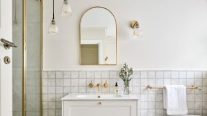 A white bathroom with a gold shower and mirror, marble tiles, and a white sink vanity with gold handles and a gold tap above it