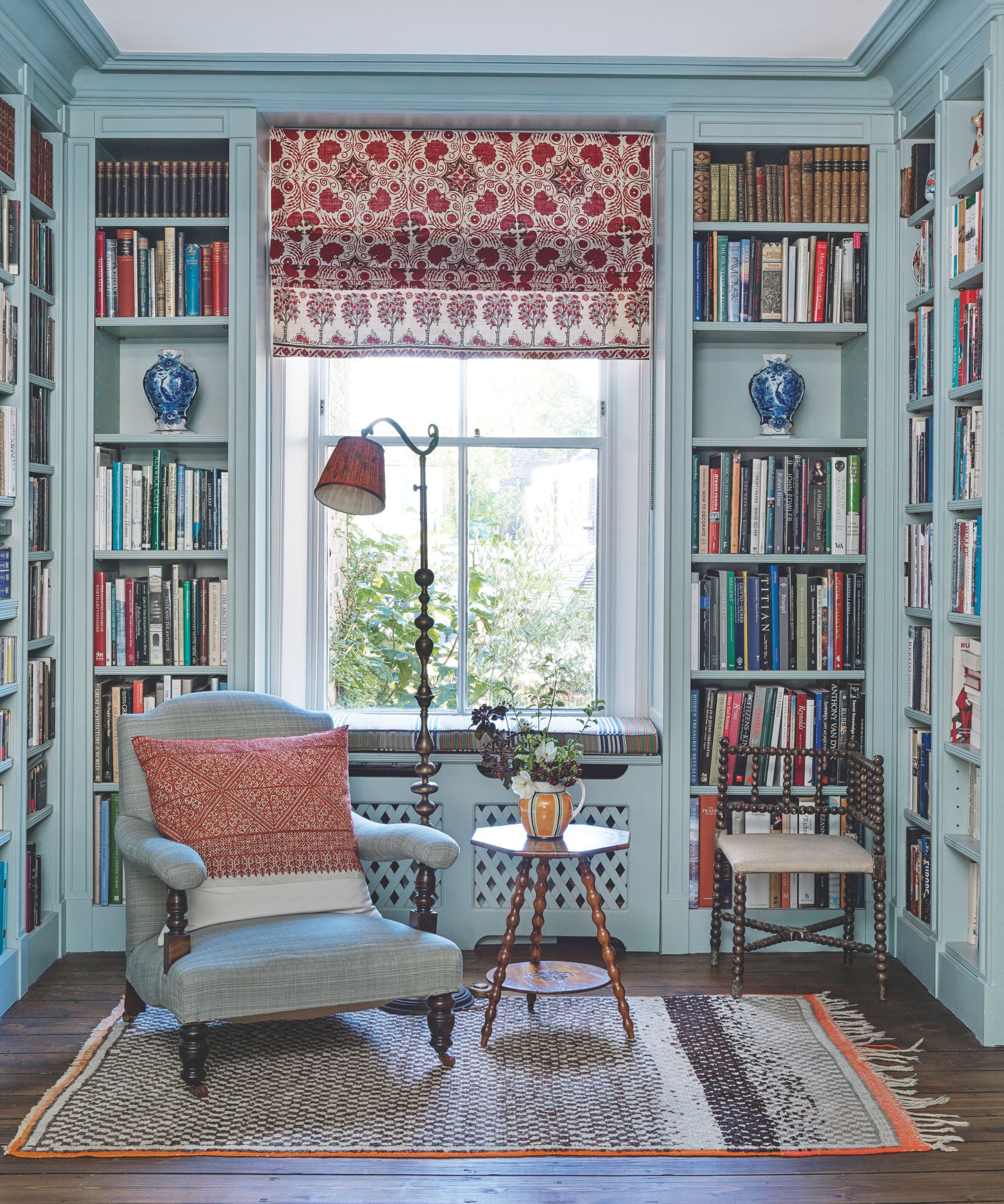 Un salon bleu avec des étagères, une chaise rembourrée bleue, un lampadaire en bois et une petite table circulaire en bois. Derrière se trouve une grande fenêtre avec un store à motifs rouges et blancs.