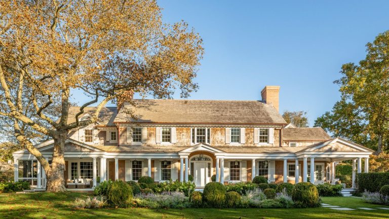 Qu’est-ce qu’une maison de style bardeau? Tout ce que vous devez savoir sur ce style architectural côtier classique Shingle style home with columns and a long porch, large tree and green lawn