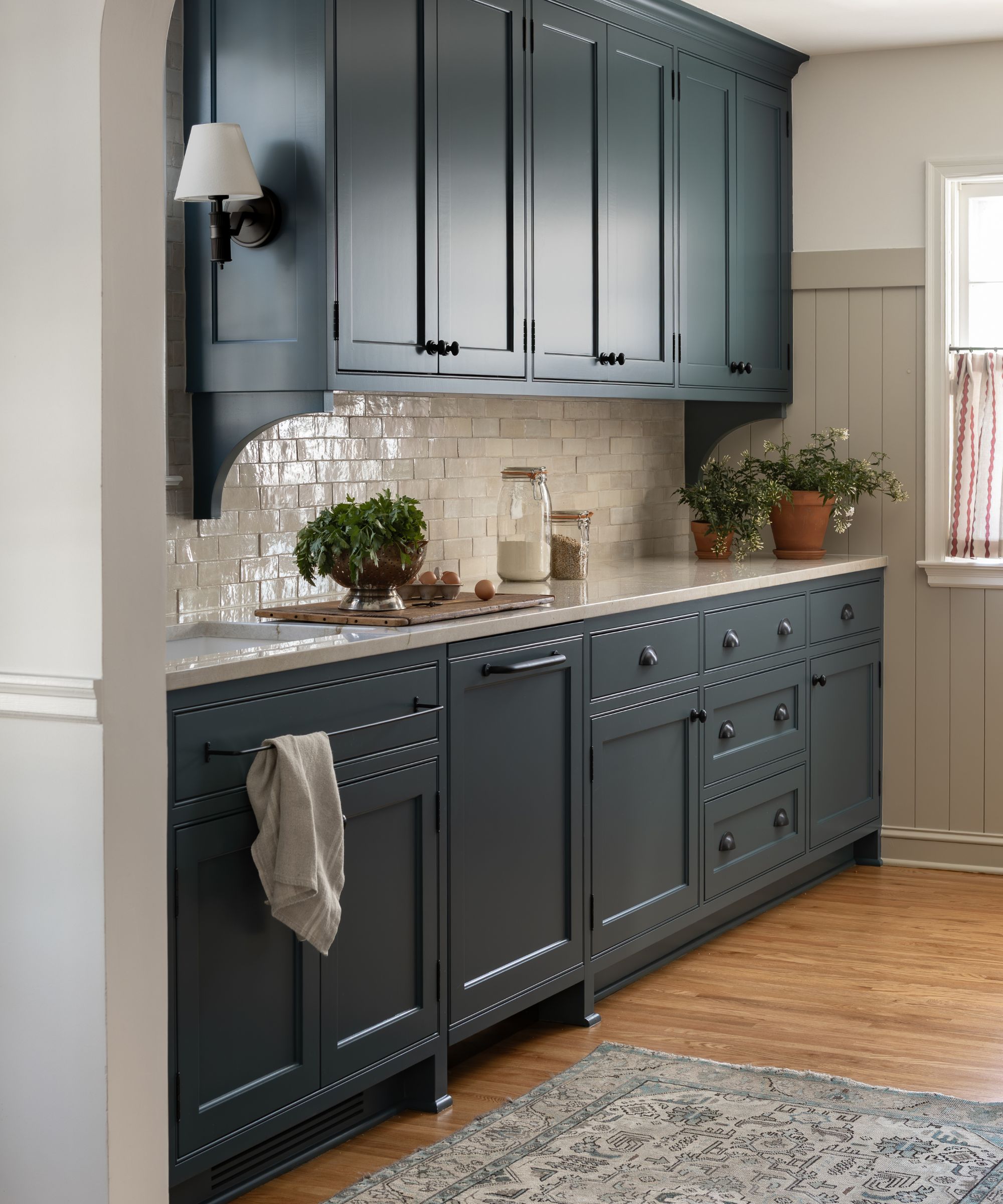 petite cuisine de cuisine bleu marine peint avec des carreaux de métro zellige blancs, un tapis gris de style vintage, des appliques murales et un rideau de café rayé