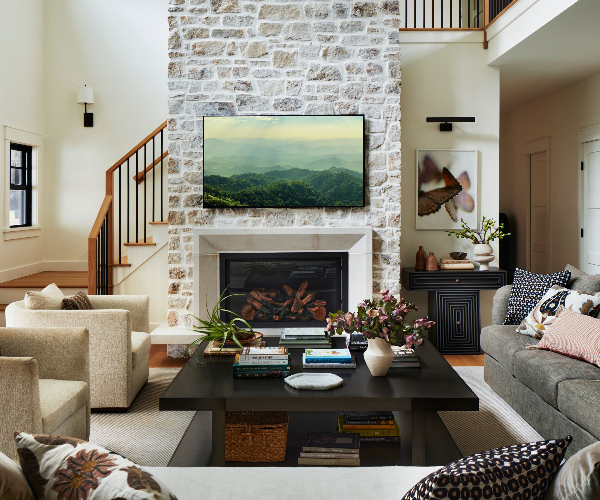 Salon ouvert de style ferme avec une grande cheminée en pierre avec cadre TV, canapés gris et beiges, fauteuils jaune beige moelleux et table basse noire