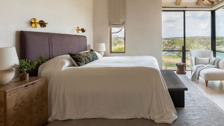 Votre chambre ressemble-t-elle à une boîte blanche ennuyeuse? Les 5 conseils faciles de ce designer d’intérieur vous aideront à le transformer en un schéma plein de personnalité en un rien de temps white bedroom with large corner windows, a white chaise lounge, a large bed with a purple upholstered headboard and a burl wood nightstand